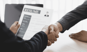 Two people shaking hands during a job interview with a resume in view.