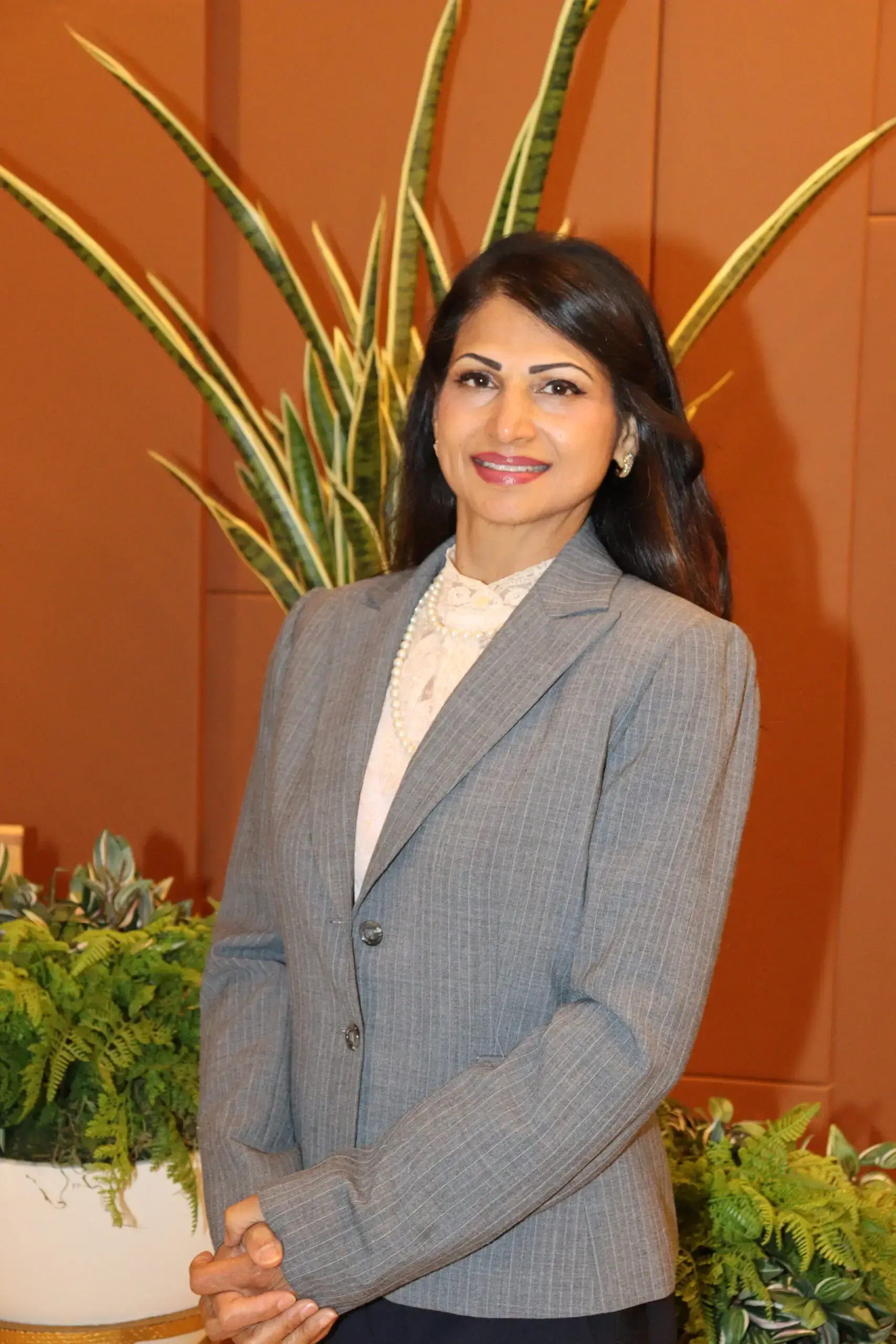 Professional woman in a gray blazer smiling indoors with plants in the background.
